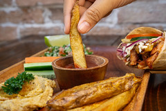Close Up Food Photo of Person using her Hands to dip French Fries in Tomato Ketchup on a Wooden Plate with Hummus, Vegetables and Doner Durum Kebab Wrap with Chicken, Tzatziki and Feta Cheese