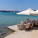 People swimming in the blue sea at Hinitsa Beach, next to restaurant tables under parasols, on a greek island