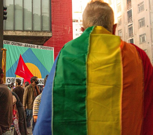 A person wearing the LGBTIQA+ flag, and in the background another flag reads “Another country is possible” on Avenida Paulista, São Paulo, Brazil. 