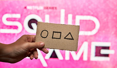 Hand holds a card with circle, square and triangle shape symbols over Squid Game logo
