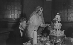 Bride cutting an elaborate wedding cake as the bridegroom looks on