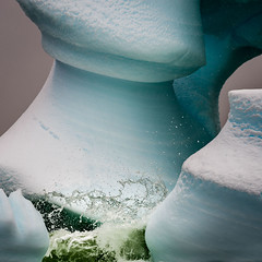 Ocean waves crash against the pillars and arches of a large iceberg sculpted by the elements.