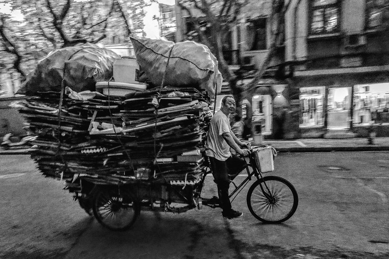 An old man engaged in recycling drives on a tricycle loaded with cardboard and other recyclables along a street, which is very dark due to the shade of the trees and the overcast weather.<br/>© <a href="https://flickr.com/people/193575245@N03" target="_blank" rel="nofollow">193575245@N03</a> (<a href="https://flickr.com/photo.gne?id=53216060257" target="_blank" rel="nofollow">Flickr</a>)