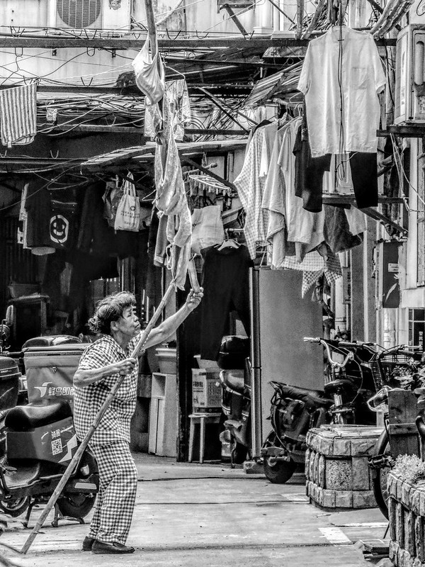 A hunchbacked old lady uses a fork pole to retrieve laundry hanging on a bamboo pole high above the alley of an old residential compound.<br/>© <a href="https://flickr.com/people/193575245@N03" target="_blank" rel="nofollow">193575245@N03</a> (<a href="https://flickr.com/photo.gne?id=53221575628" target="_blank" rel="nofollow">Flickr</a>)