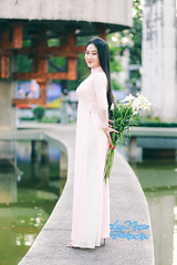 In delicate light-pink traditional Vietnamese dress, a graceful maiden holds a bunch of white calla lilies, posing on a curved structure on a green lake at a street intersection. The soft afternoon sunlight and pale blue smoke evoke an ancient ambiance.