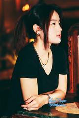 A beautiful girl stands at a wooden table in the corner of a room filled with nostalgic items, with a soft, dim light from an electric lamp in the background. The photo uses the warm, rich tones of Kodak Gold 200 film, giving it a classic and vintage feel