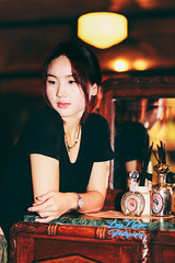 A beautiful girl stands at a wooden table in the corner of a room filled with nostalgic items, with a soft, dim light from an electric lamp in the background. The photo uses the warm, rich tones of Kodak Gold 200 film, giving it a classic and vintage feel